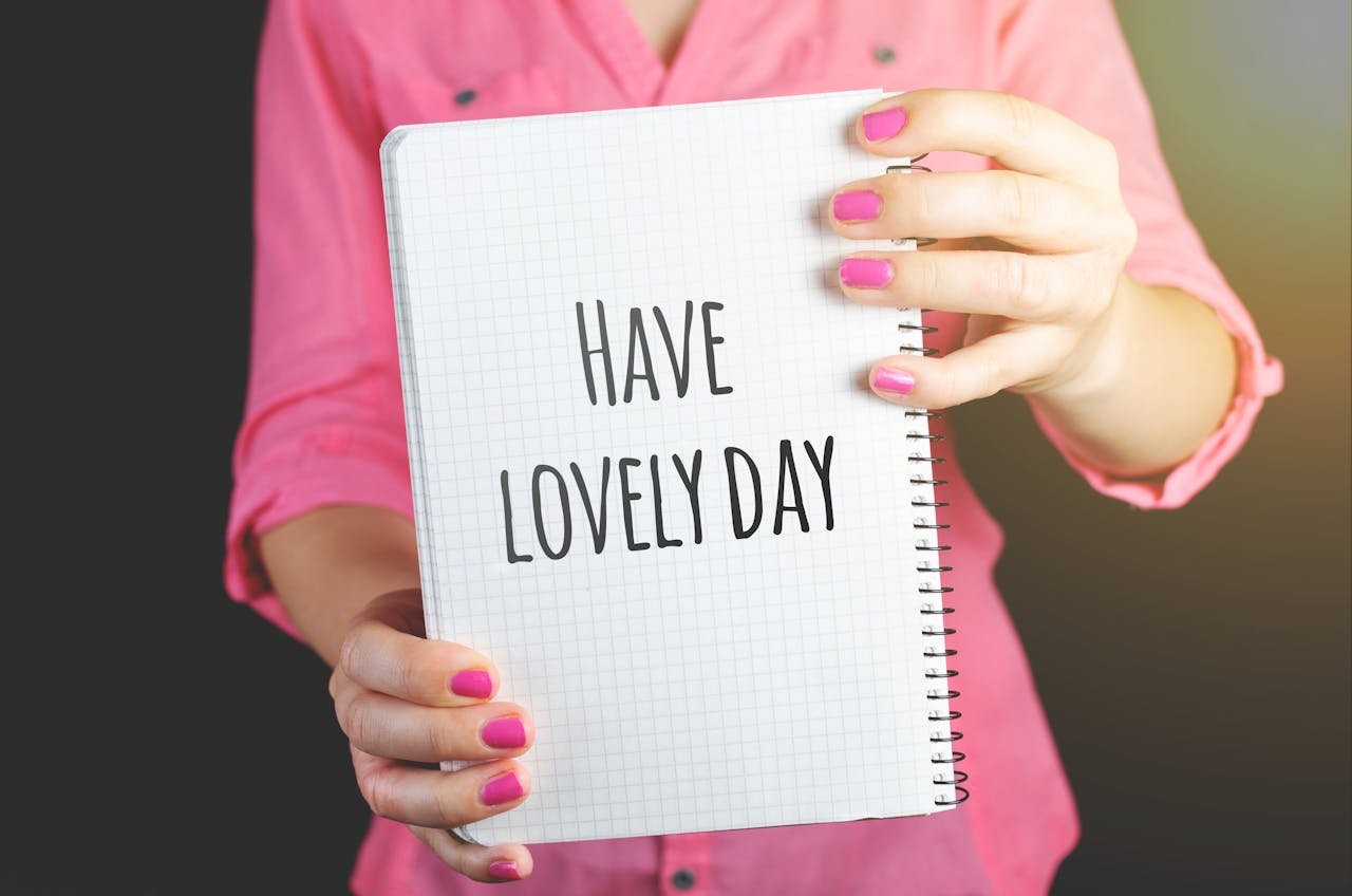 A woman in a pink shirt holds a notebook displaying 'Have a Lovely Day', conveying positivity.